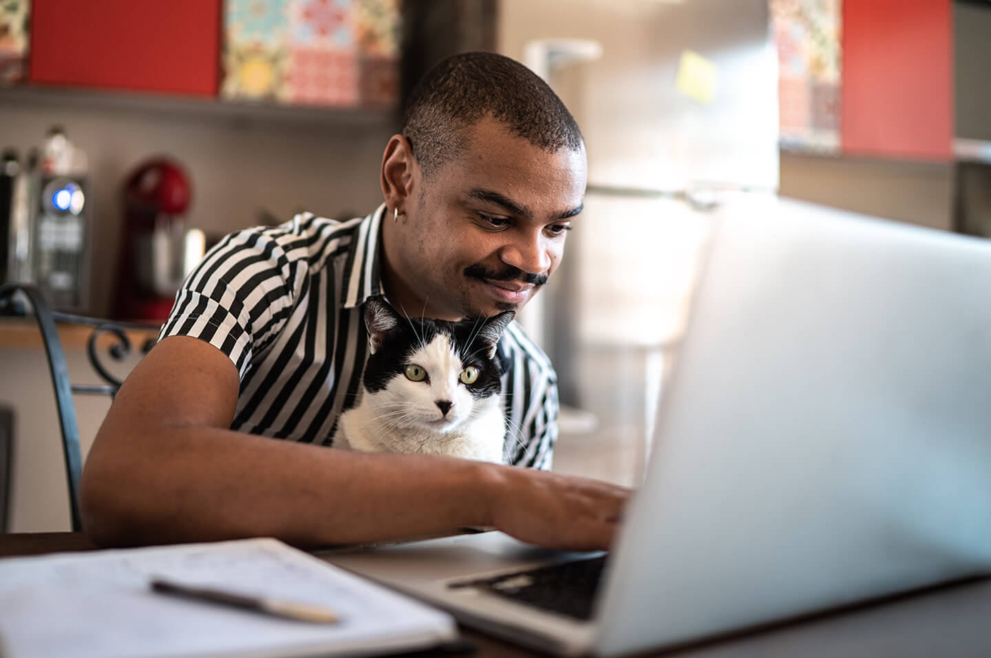 Man sitting at their kitchen table using a laptop with a cat on their lap. Credit: FG Trade